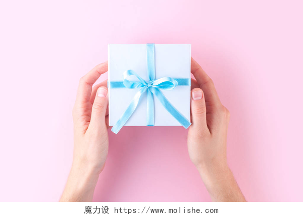 在粉色的桌子上一个人的双手拿着白色的礼盒女性手拿着一个白色的小礼品盒包裹着蓝色的丝带在粉红色的背景。给予和接受来自亲人的礼物. 